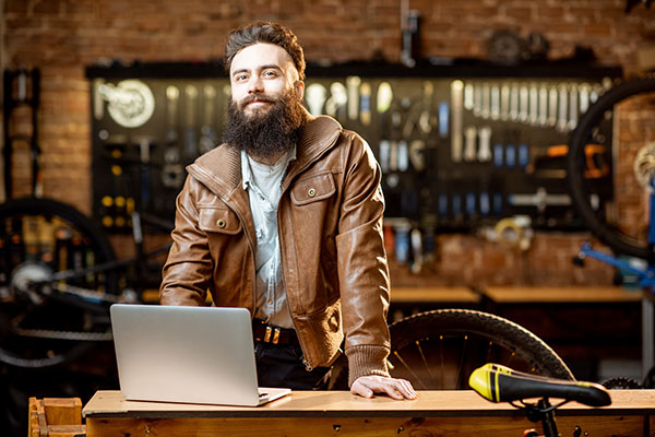 Portrait of a man in the bicycle store | Portrait of a handsome bearded man as bicycle store owner or manager standing with laptop at the bicycle workshop